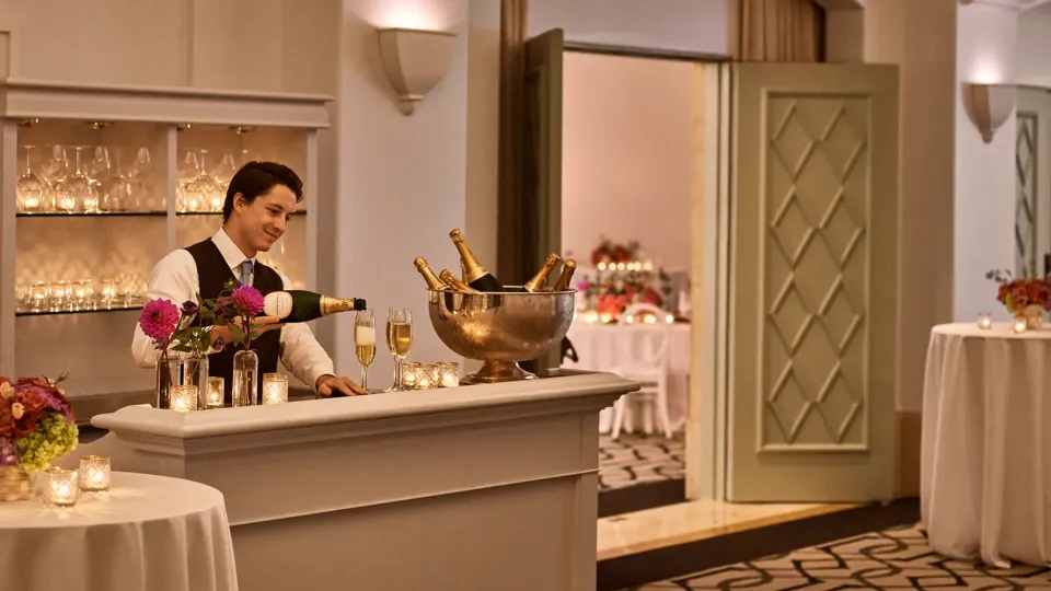 Ballroom foyer bar with bartender pouring champagne, votive candles, and floral décor in warm ambient lighting.
