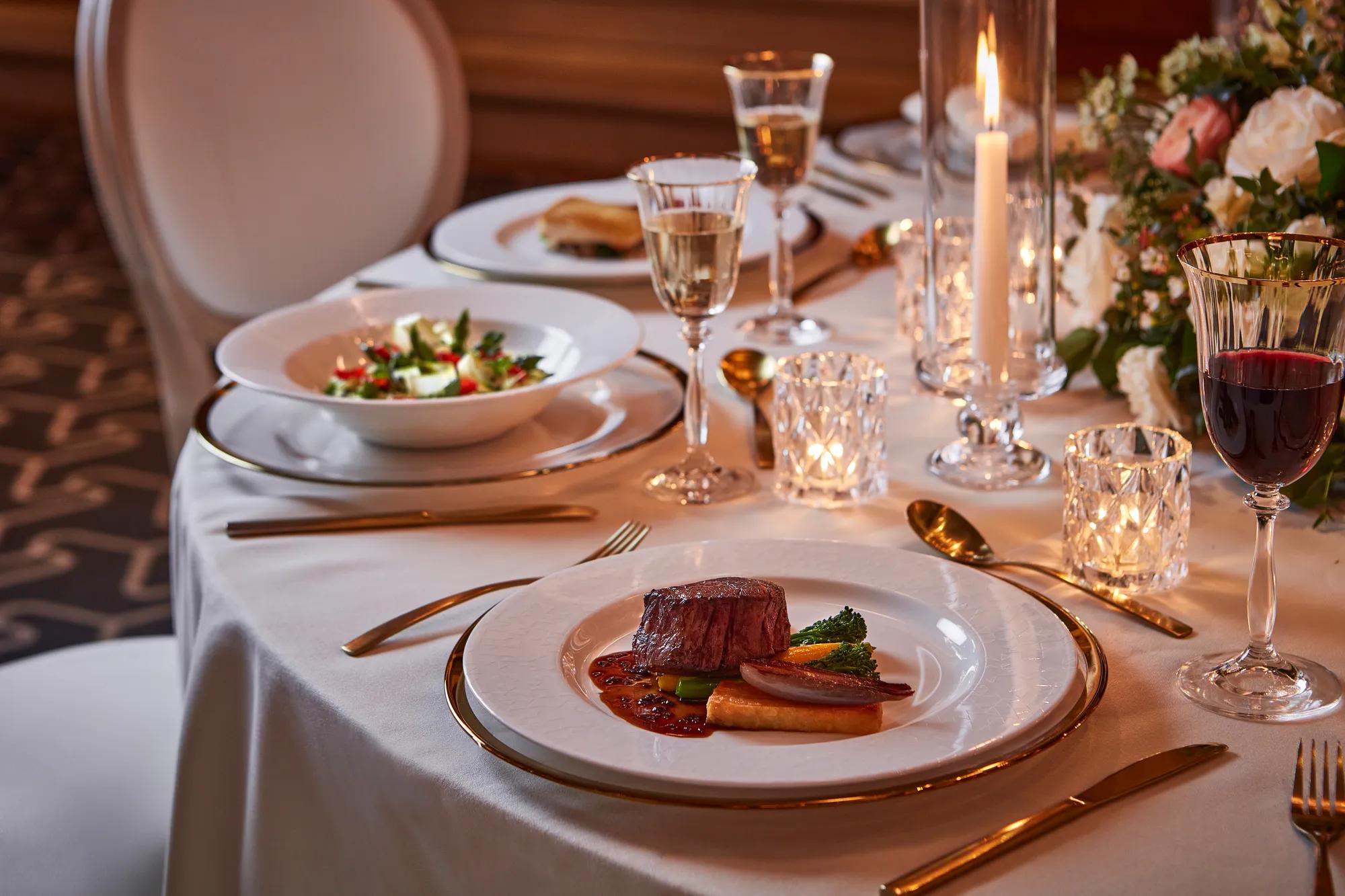 Close-up of plated beef dish with vegetables, salad, wine glasses, and candlelight on white tablecloth.
