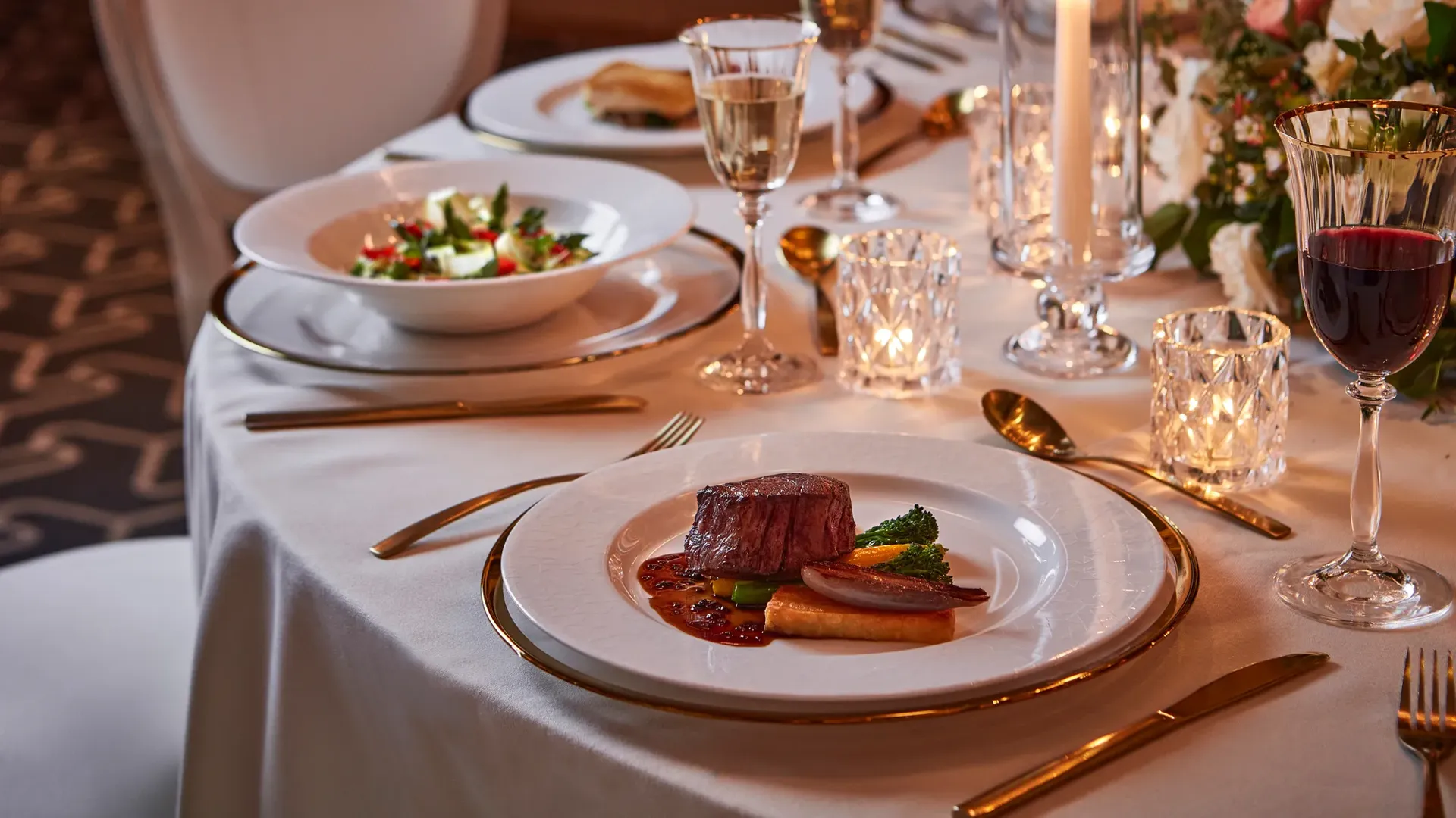 Close-up of plated beef dish with vegetables, salad, wine glasses, and candlelight on white tablecloth.