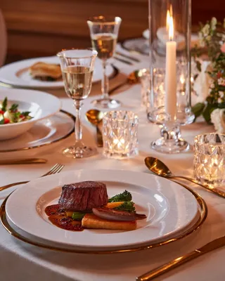 Close-up of plated beef dish with vegetables, salad, wine glasses, and candlelight on white tablecloth.