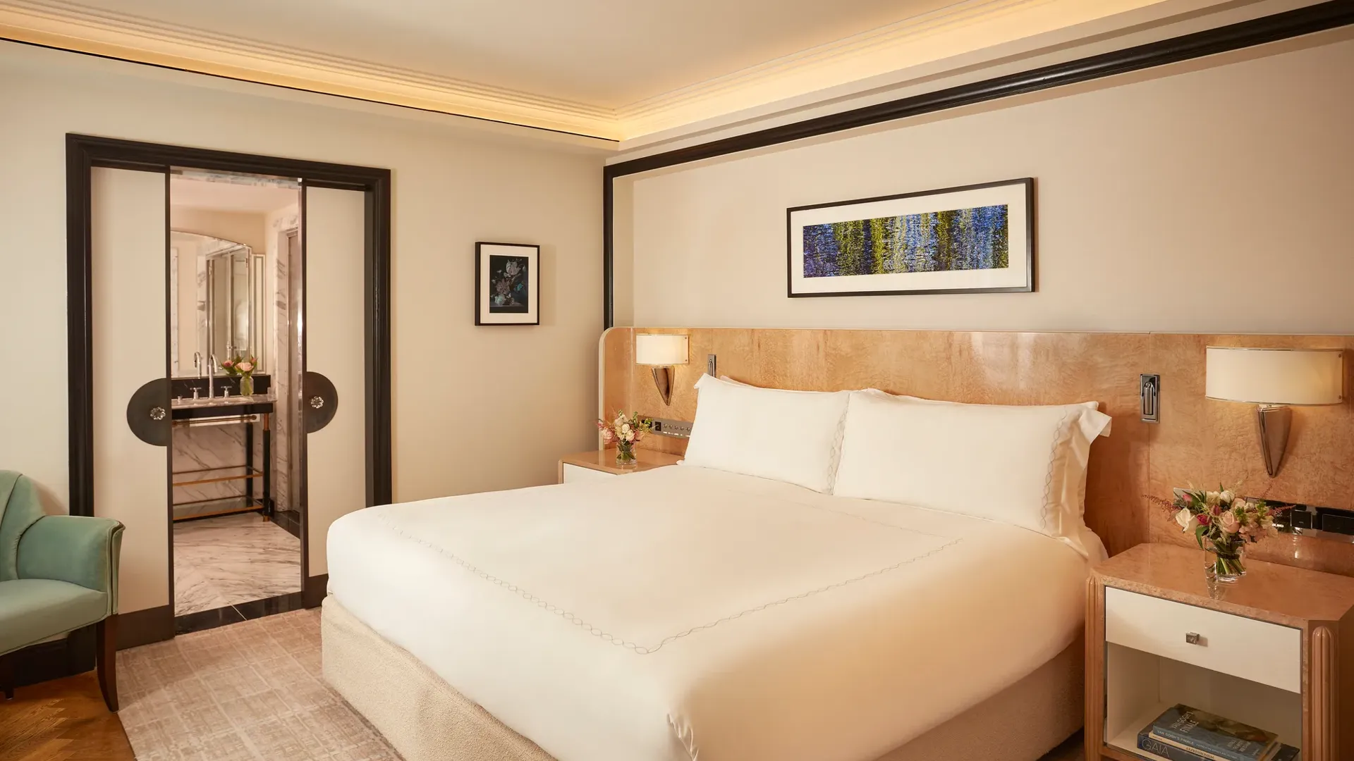 Elegant bedroom featuring a king-size bed with crisp white linens, light wood bedside tables with flowers, and a framed artwork above the headboard, with sliding doors opening to a marble bathroom.