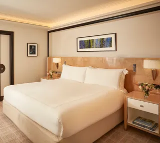 Elegant bedroom featuring a king-size bed with crisp white linens, light wood bedside tables with flowers, and a framed artwork above the headboard, with sliding doors opening to a marble bathroom.