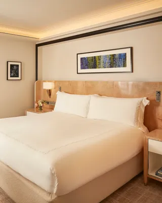 Elegant bedroom featuring a king-size bed with crisp white linens, light wood bedside tables with flowers, and a framed artwork above the headboard, with sliding doors opening to a marble bathroom.