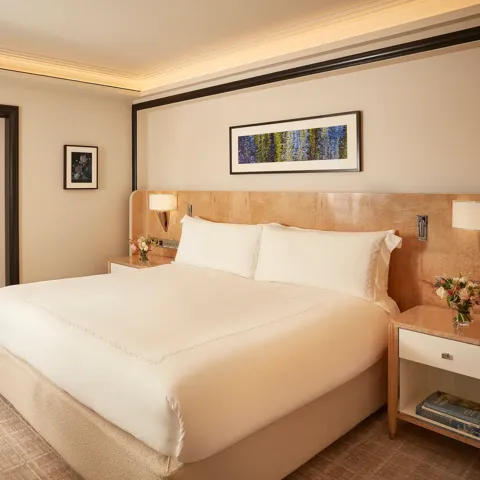 Elegant bedroom featuring a king-size bed with crisp white linens, light wood bedside tables with flowers, and a framed artwork above the headboard, with sliding doors opening to a marble bathroom.