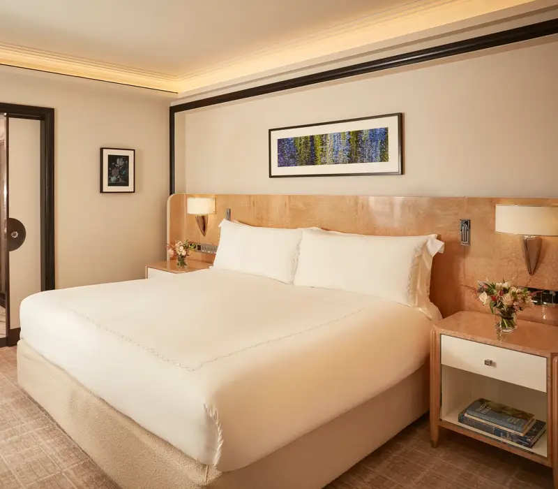Elegant bedroom featuring a king-size bed with crisp white linens, light wood bedside tables with flowers, and a framed artwork above the headboard, with sliding doors opening to a marble bathroom.