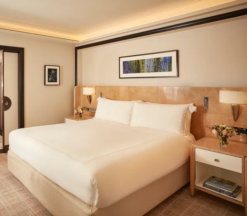 Elegant bedroom featuring a king-size bed with crisp white linens, light wood bedside tables with flowers, and a framed artwork above the headboard, with sliding doors opening to a marble bathroom.
