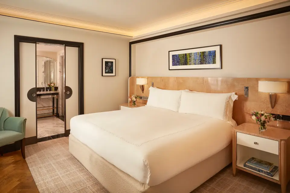 Elegant bedroom featuring a king-size bed with crisp white linens, light wood bedside tables with flowers, and a framed artwork above the headboard, with sliding doors opening to a marble bathroom.