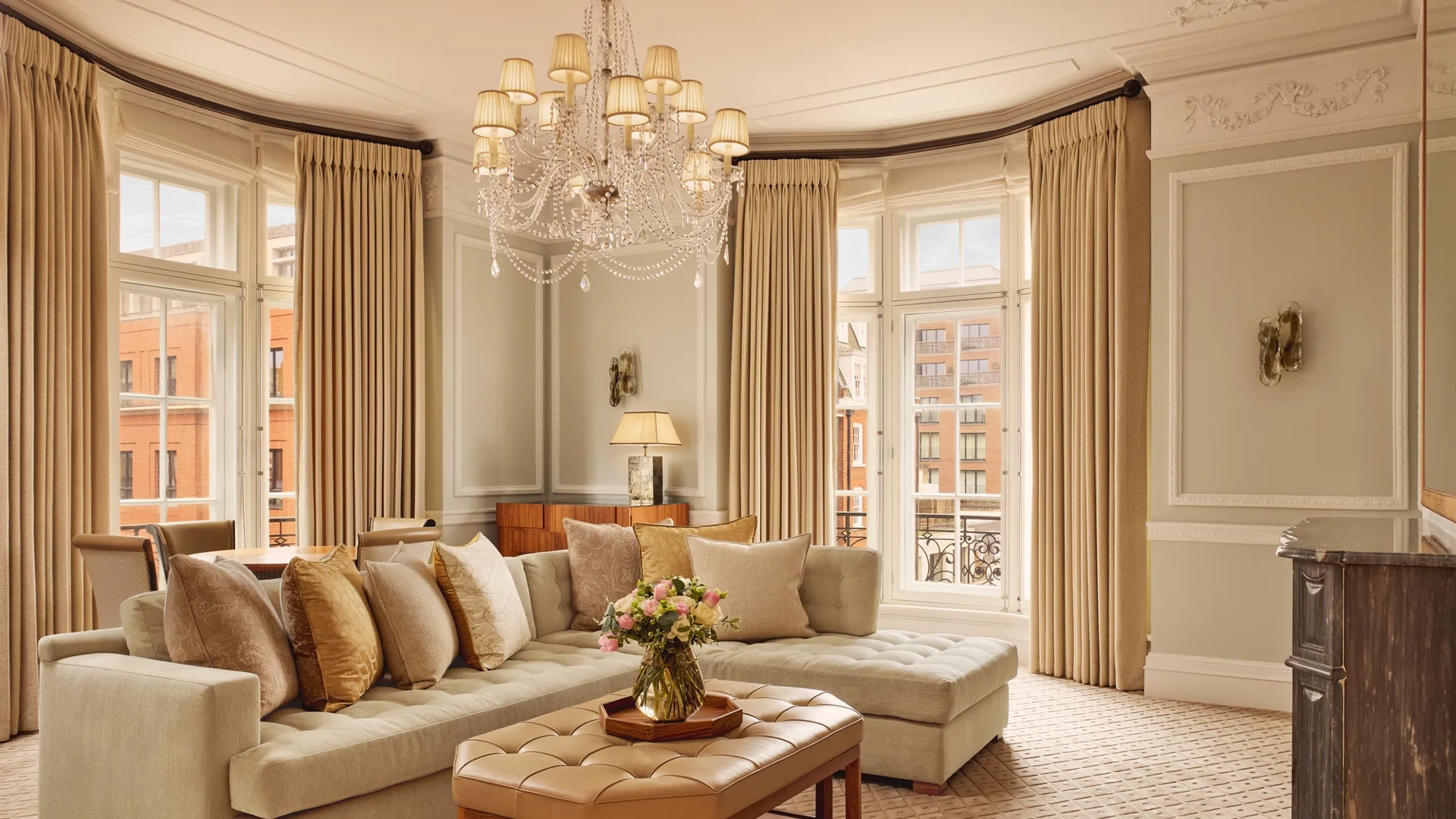 Elegant living room with a large cream corner sofa, tufted leather coffee table, and chandelier, surrounded by tall windows with beige curtains and city views beyond.