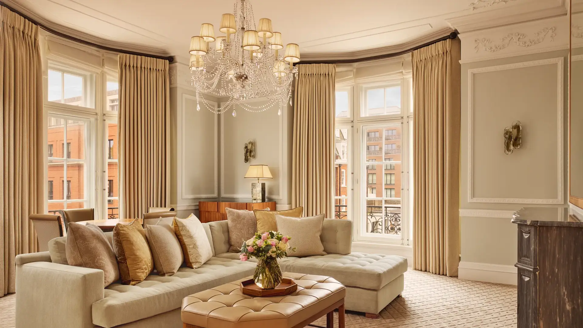 Elegant living room with a large cream corner sofa, tufted leather coffee table, and chandelier, surrounded by tall windows with beige curtains and city views beyond.