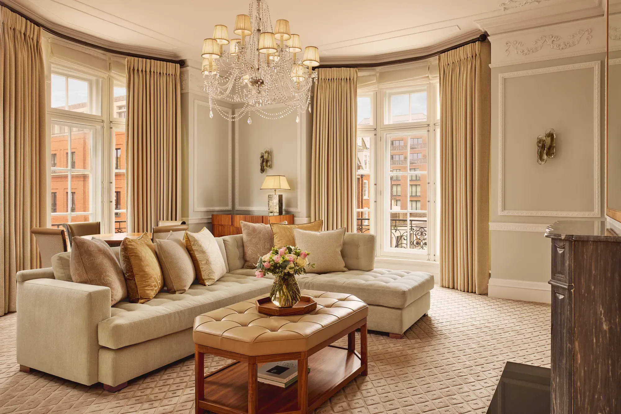 Elegant living room with a large cream corner sofa, tufted leather coffee table, and chandelier, surrounded by tall windows with beige curtains and city views beyond.