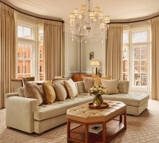 Elegant living room with a large cream corner sofa, tufted leather coffee table, and chandelier, surrounded by tall windows with beige curtains and city views beyond.