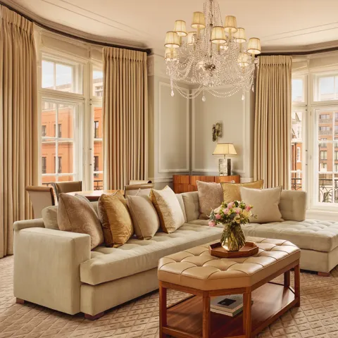 Elegant living room with a large cream corner sofa, tufted leather coffee table, and chandelier, surrounded by tall windows with beige curtains and city views beyond.