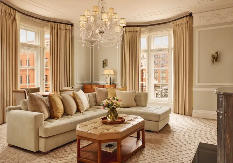 Elegant living room with a large cream corner sofa, tufted leather coffee table, and chandelier, surrounded by tall windows with beige curtains and city views beyond.