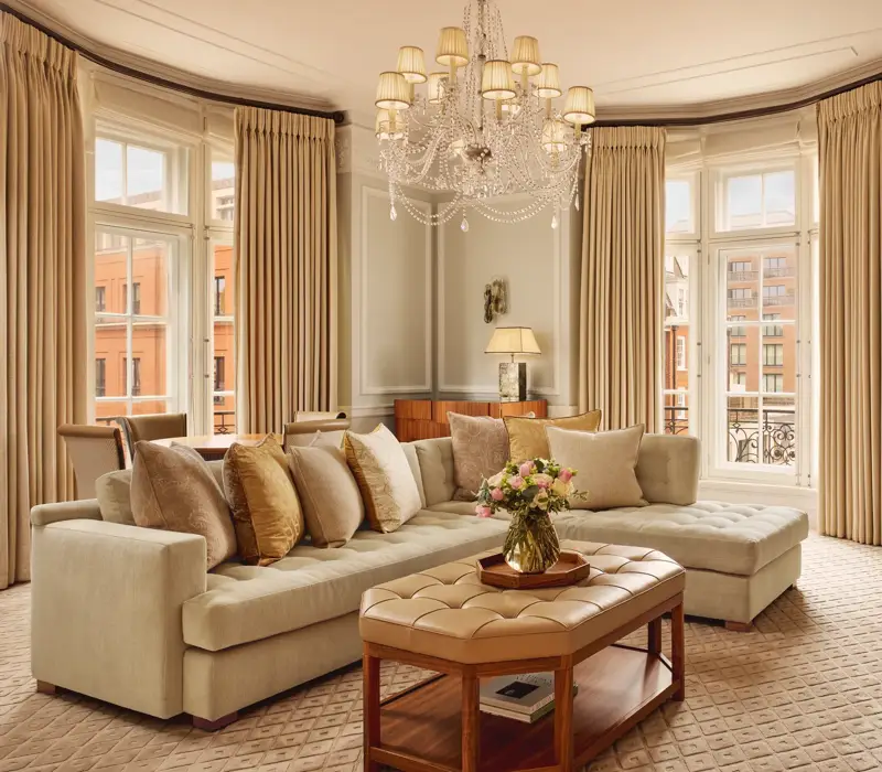 Elegant living room with a large cream corner sofa, tufted leather coffee table, and chandelier, surrounded by tall windows with beige curtains and city views beyond.