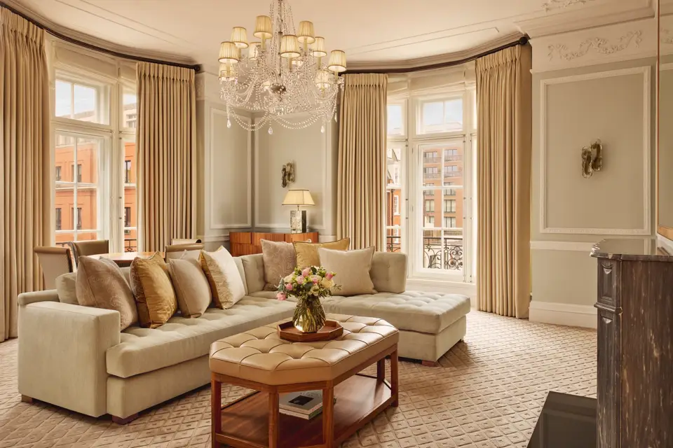 Elegant living room with a large cream corner sofa, tufted leather coffee table, and chandelier, surrounded by tall windows with beige curtains and city views beyond.