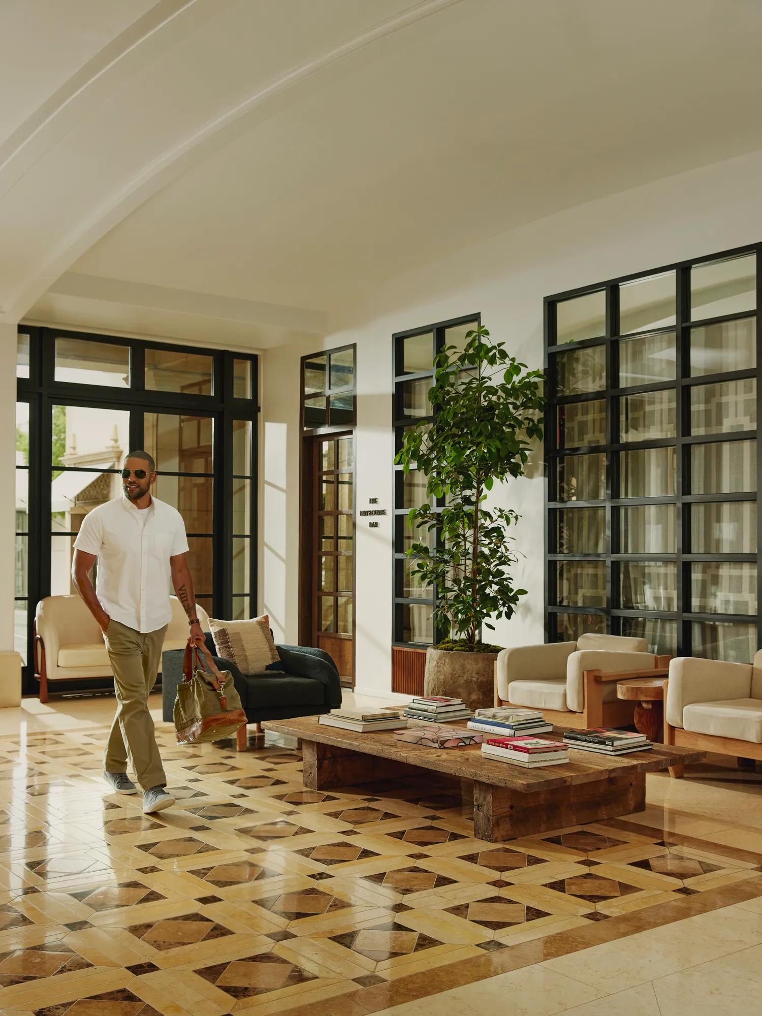 Person carrying a travel bag walks through a bright hotel lobby with patterned tile floors, modern seating, large windows, and indoor plants.