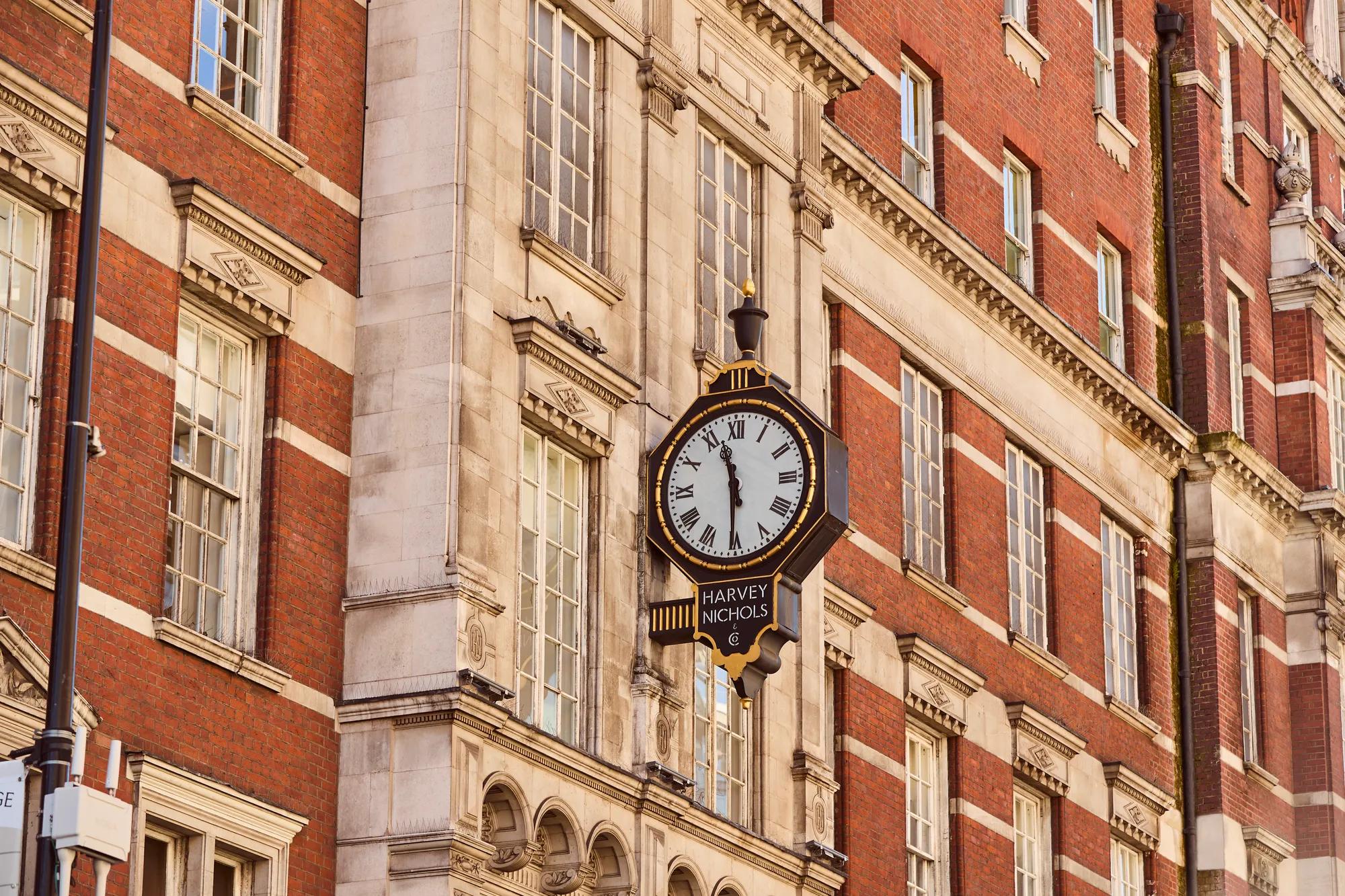 Harvey Nichols clock mounted on a red-brick building facade with tall windows and decorative stone architectural details.