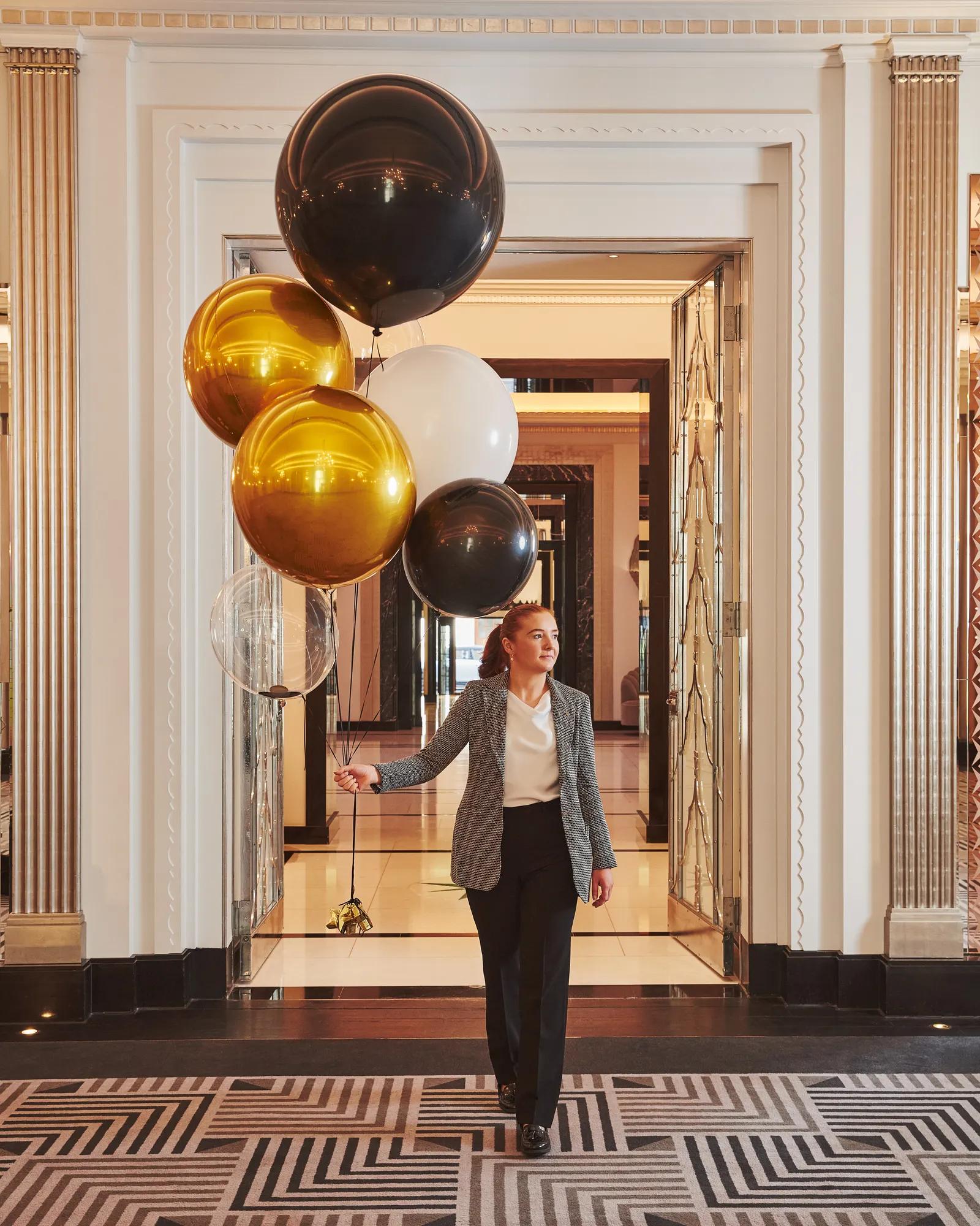 Guest walking through an elegant hotel corridor holding black, gold, and white balloons beneath decorative archways.