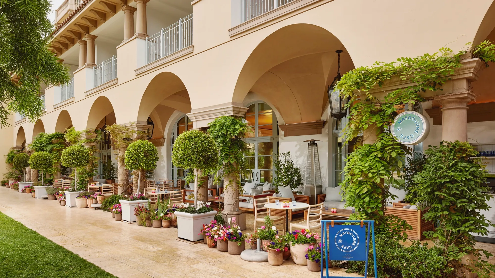 Arched outdoor café terrace lined with topiaries, vines, flowers, and a blue “Maybourne Café” sign.