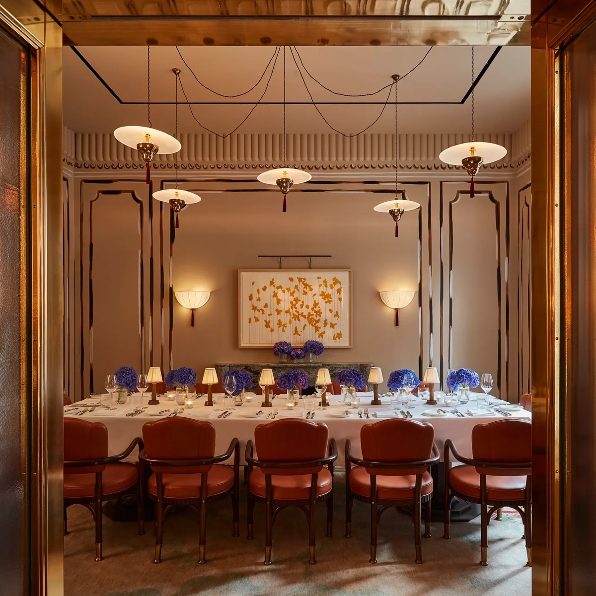 Formal dining room with long table, red upholstered chairs, blue floral centrepieces, and warm pendant lighting viewed through open doors.