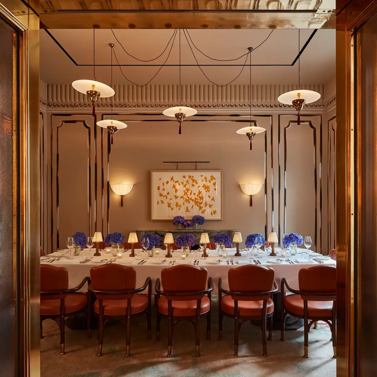 Formal dining room with long table, red upholstered chairs, blue floral centrepieces, and warm pendant lighting viewed through open doors.