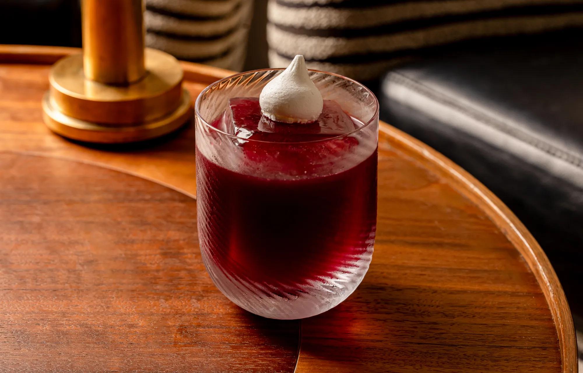 Deep red cocktail with foam garnish in textured glass on wooden table.