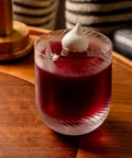 Deep red cocktail with foam garnish in textured glass on wooden table.