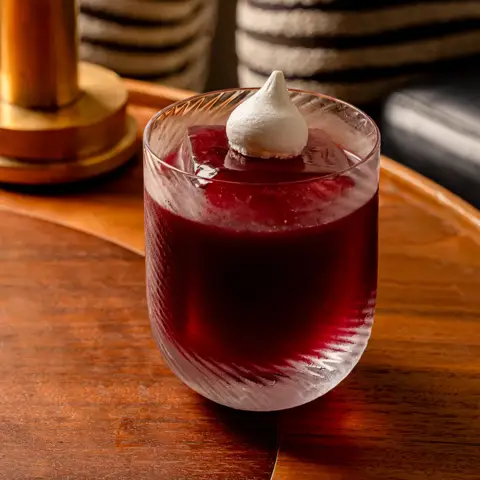Deep red cocktail with foam garnish in textured glass on wooden table.