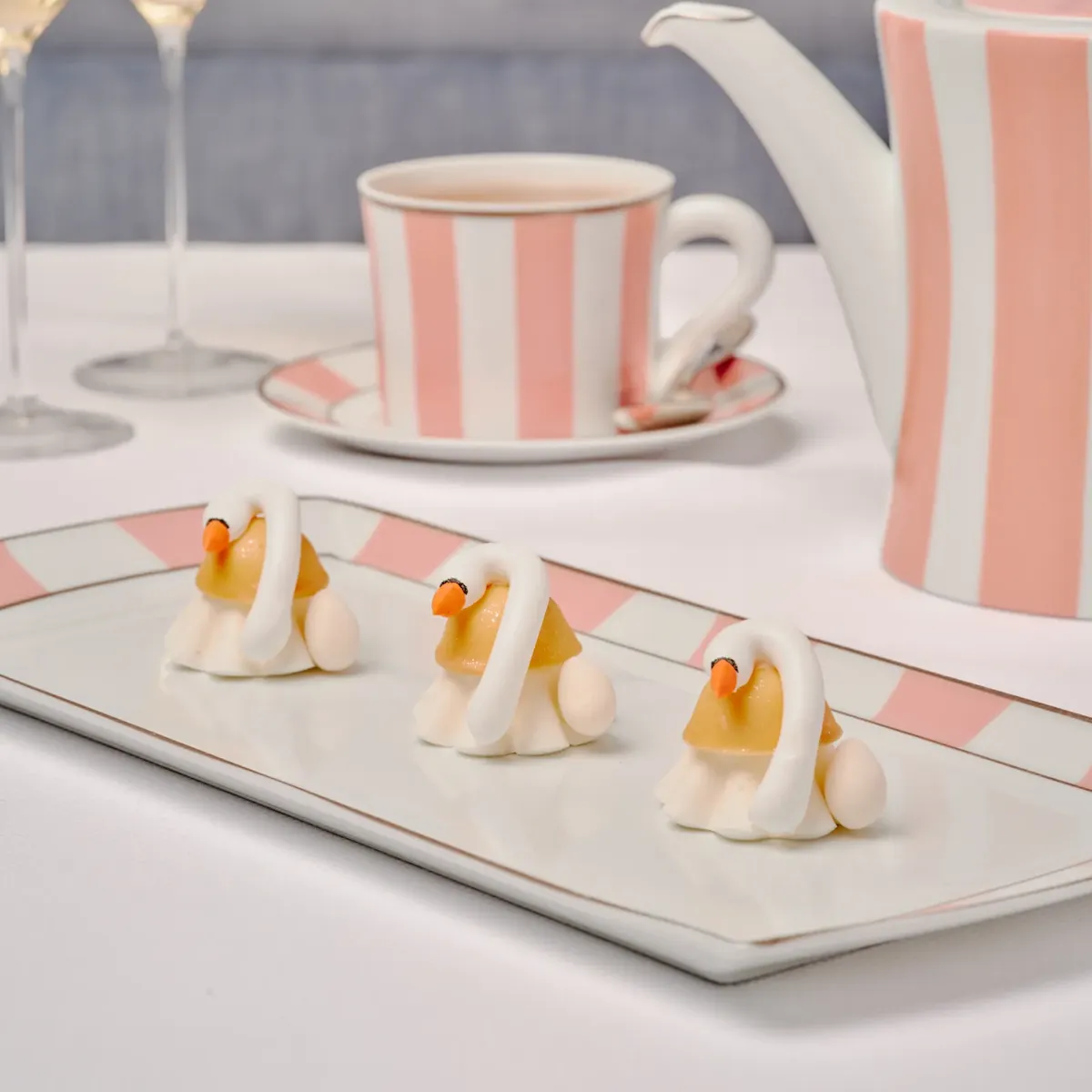 Three swan-shaped petits fours on a tray with champagne and pink-striped teacup and teapot.