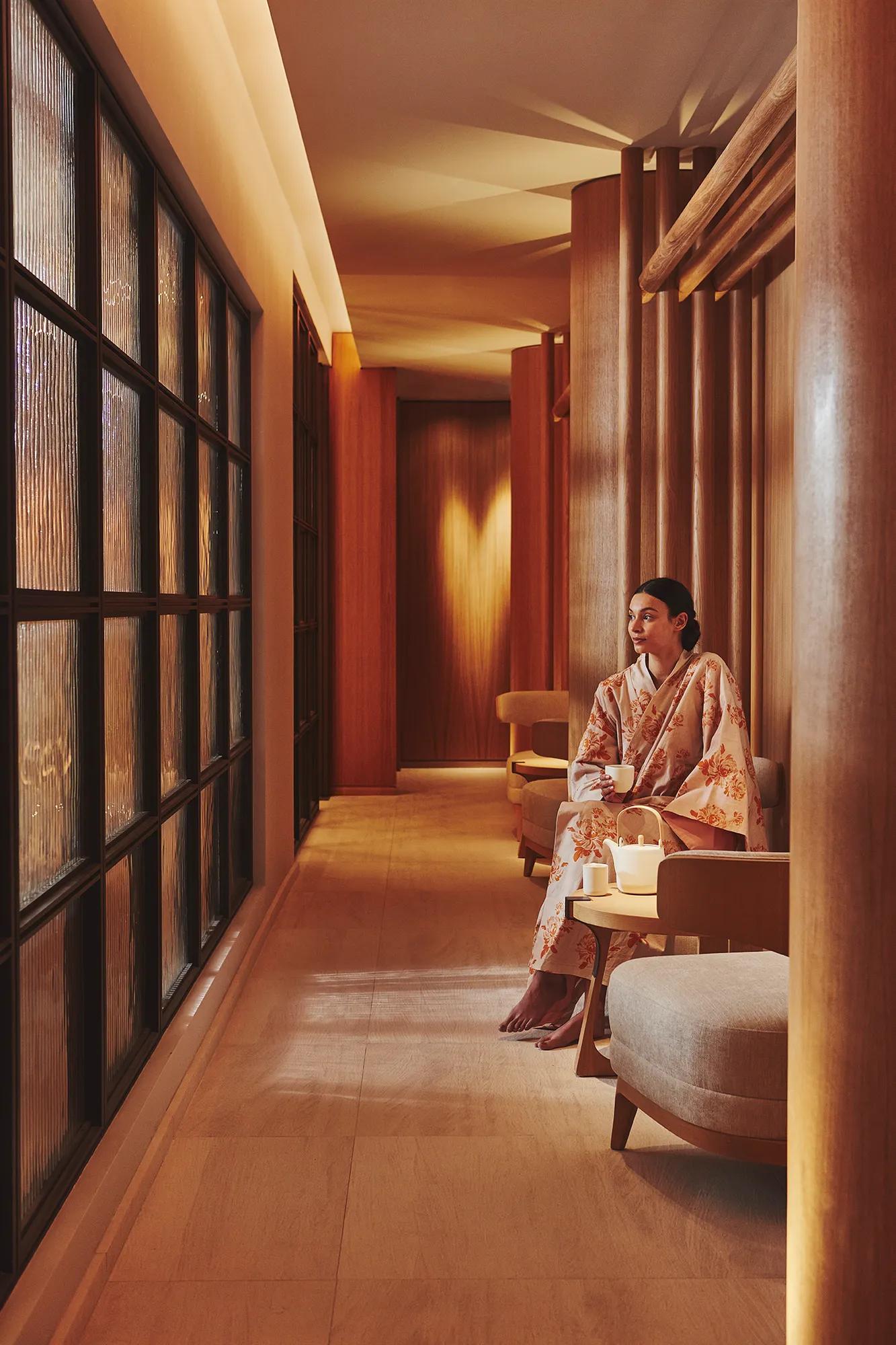A woman in a floral kimono sits in a softly lit spa corridor at The Maybourne Riviera, beside a frosted glass wall and warm wooden interiors.
