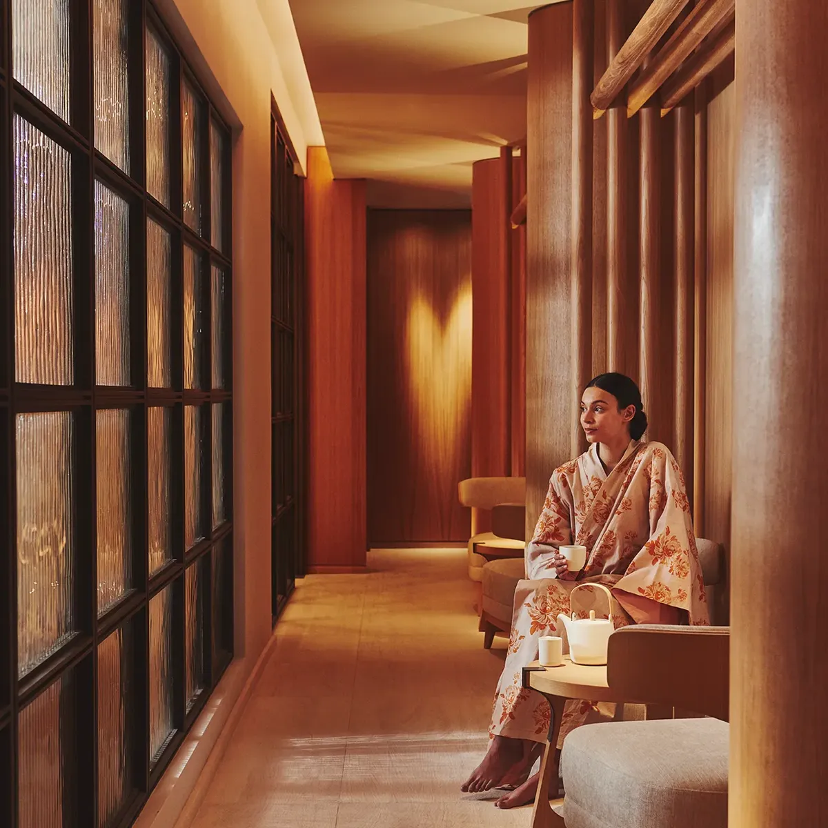 A woman in a floral kimono sits in a softly lit spa corridor at The Maybourne Riviera, beside a frosted glass wall and warm wooden interiors.