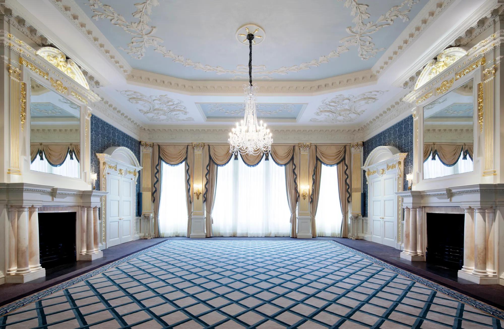 Grand Claridge’s ballroom with ornate white and gold detailing, a central crystal chandelier, tall curtained windows, and mirrored fireplaces on either side of the room.