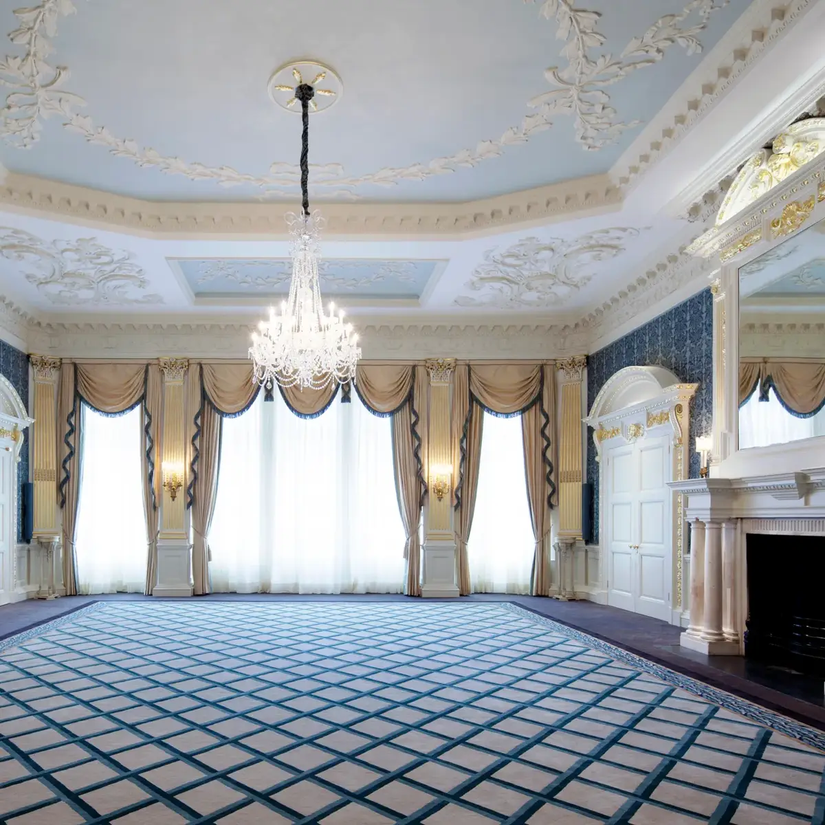 Grand Claridge’s ballroom with ornate white and gold detailing, a central crystal chandelier, tall curtained windows, and mirrored fireplaces on either side of the room.
