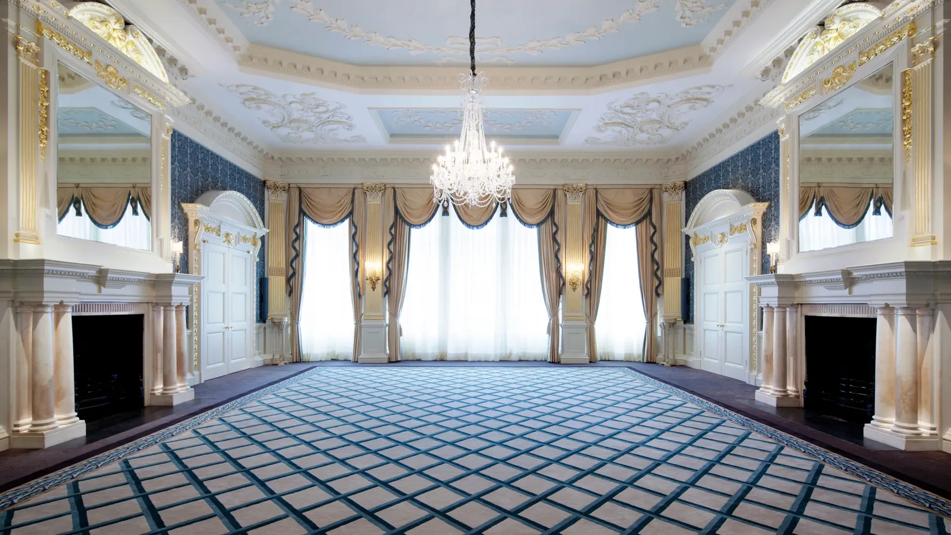 Grand Claridge’s ballroom with ornate white and gold detailing, a central crystal chandelier, tall curtained windows, and mirrored fireplaces on either side of the room.