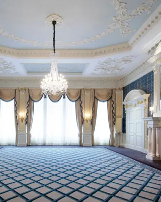 Grand Claridge’s ballroom with ornate white and gold detailing, a central crystal chandelier, tall curtained windows, and mirrored fireplaces on either side of the room.