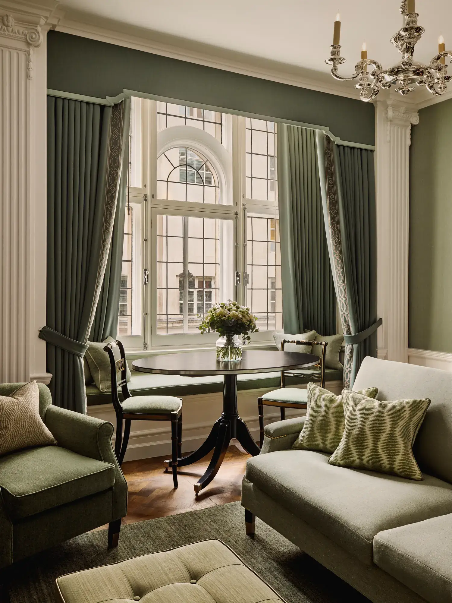 Sitting area with green sofa and armchairs, round dining table, large arched windows, and elegant draped curtains.