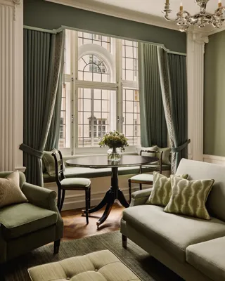 Sitting area with green sofa and armchairs, round dining table, large arched windows, and elegant draped curtains.