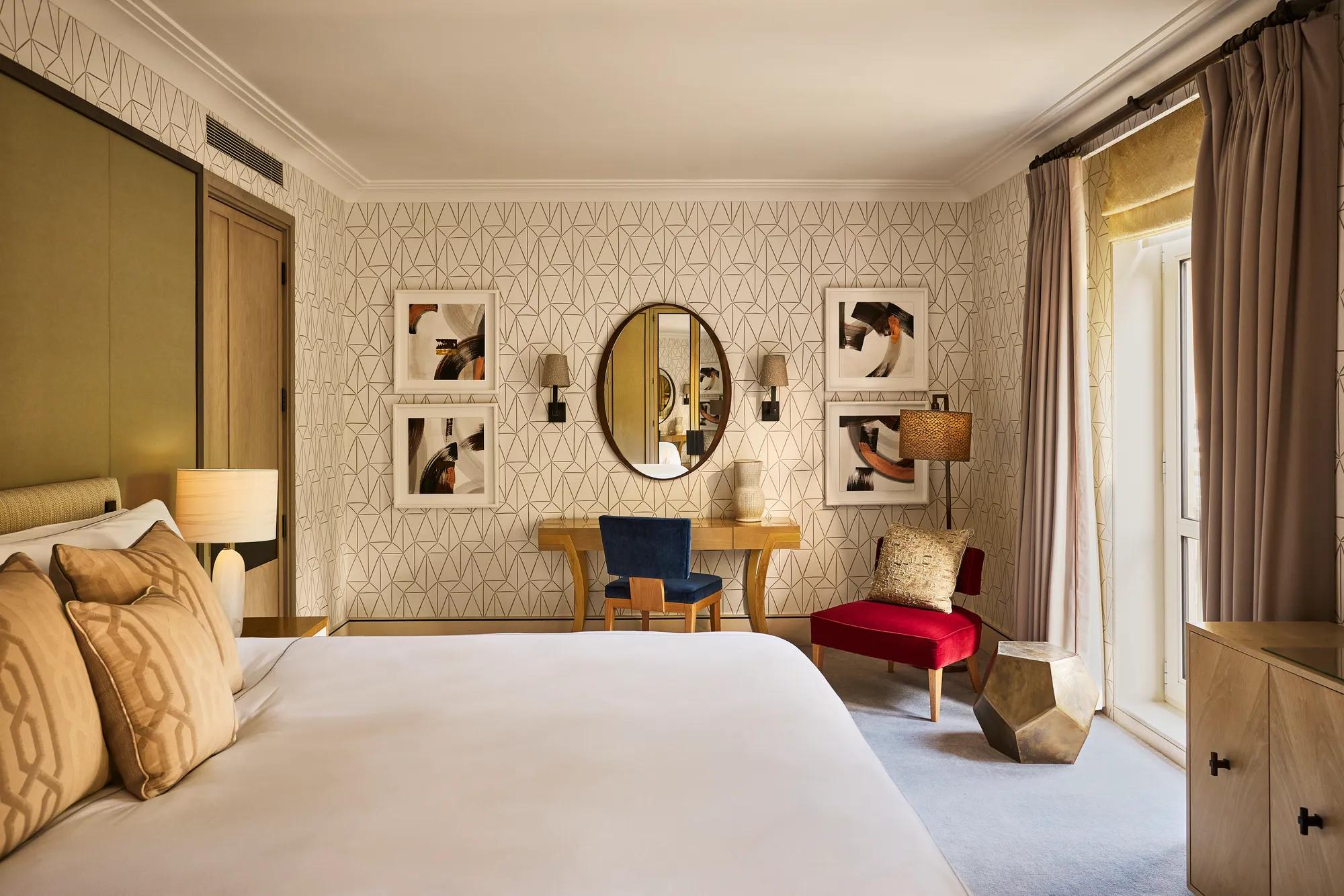 Bedroom with patterned wallpaper, desk and mirror, blue and red accent chairs, and French doors.