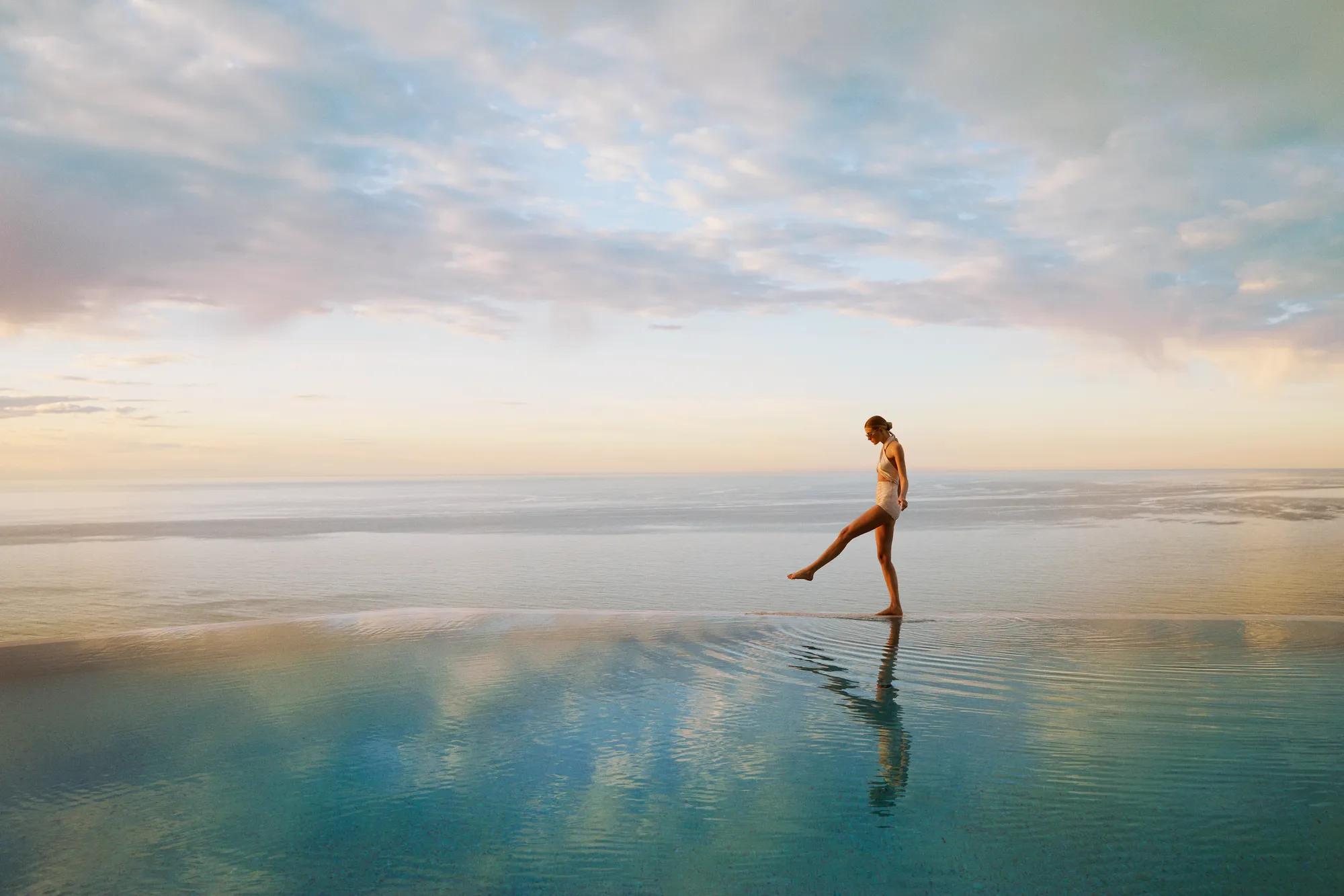 Person walking across a calm infinity pool at sunrise, with ocean horizon and soft pastel sky reflected in water.