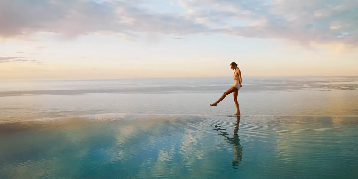 Person walking across a calm infinity pool at sunrise, with ocean horizon and soft pastel sky reflected in water.