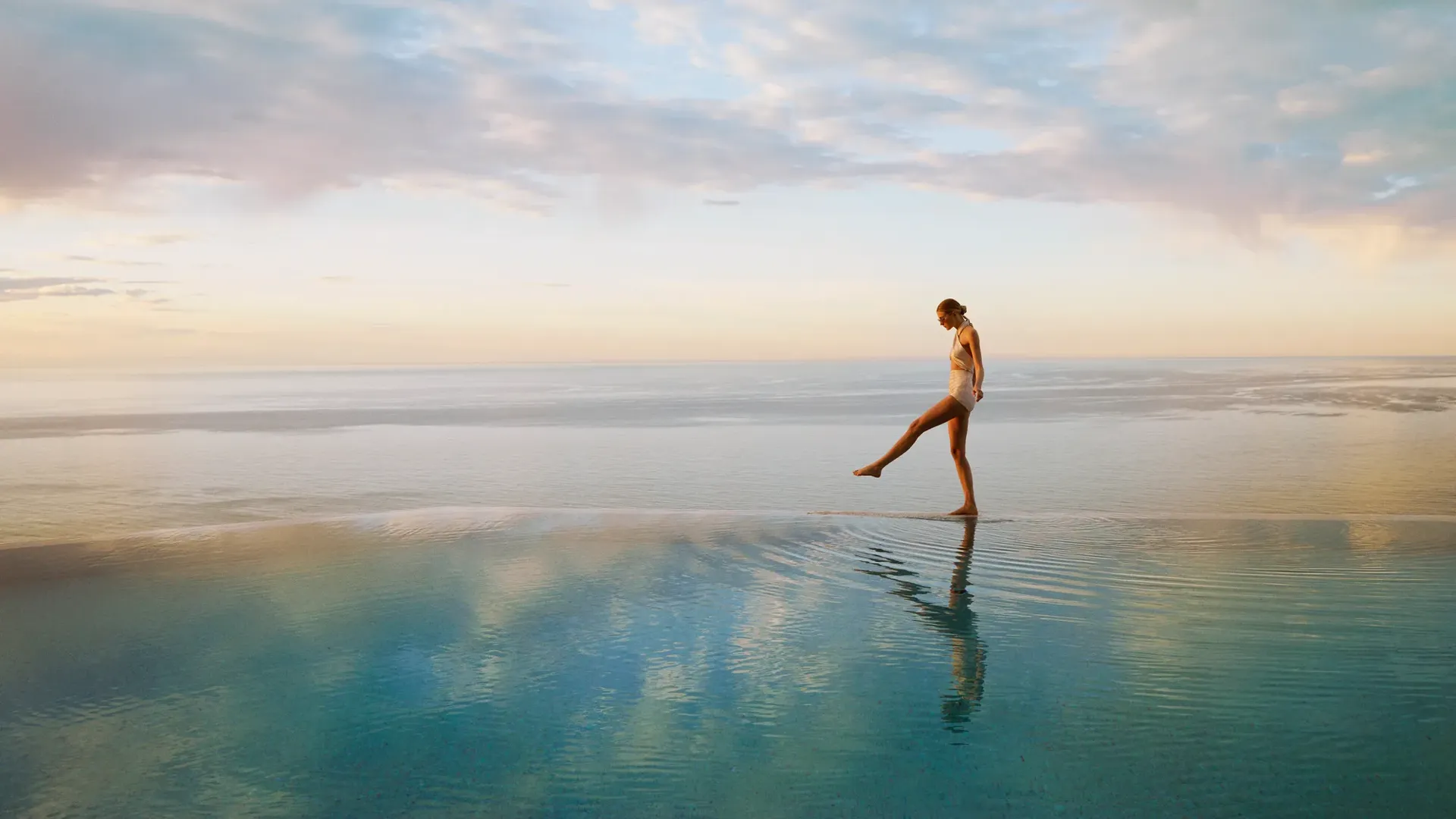 Person walking across a calm infinity pool at sunrise, with ocean horizon and soft pastel sky reflected in water.
