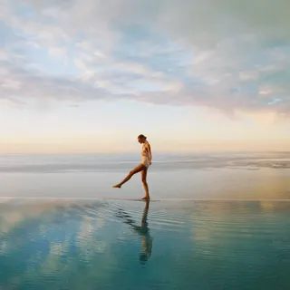 Person walking across a calm infinity pool at sunrise, with ocean horizon and soft pastel sky reflected in water.