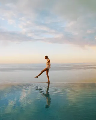 Person walking across a calm infinity pool at sunrise, with ocean horizon and soft pastel sky reflected in water.