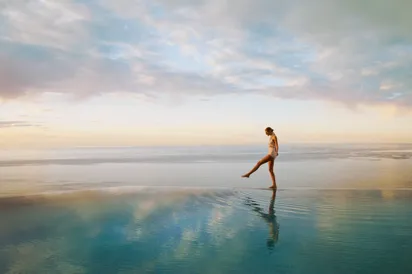 Person walking across a calm infinity pool at sunrise, with ocean horizon and soft pastel sky reflected in water.