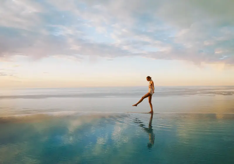 Person walking across a calm infinity pool at sunrise, with ocean horizon and soft pastel sky reflected in water.