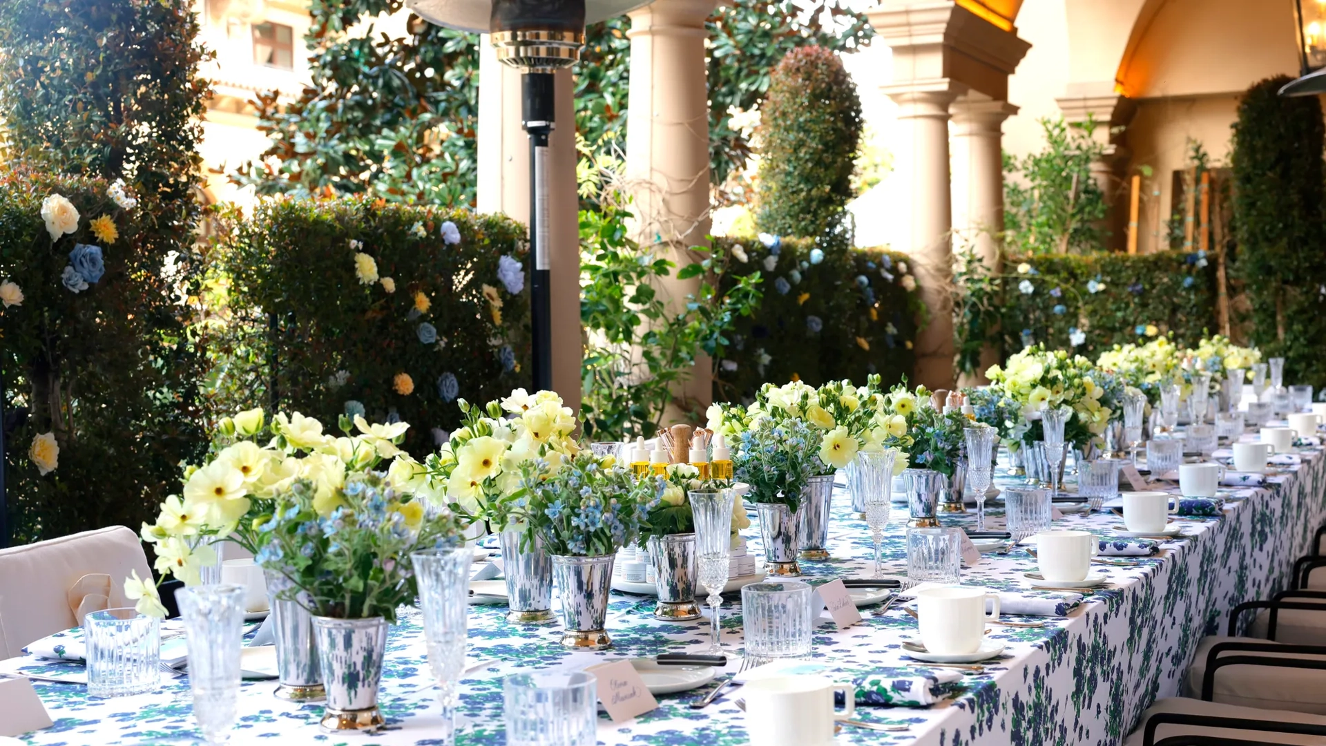Kitchen Patio long table dressed in blue-floral linens with pastel centerpieces beneath an arched colonnade and garden hedges.