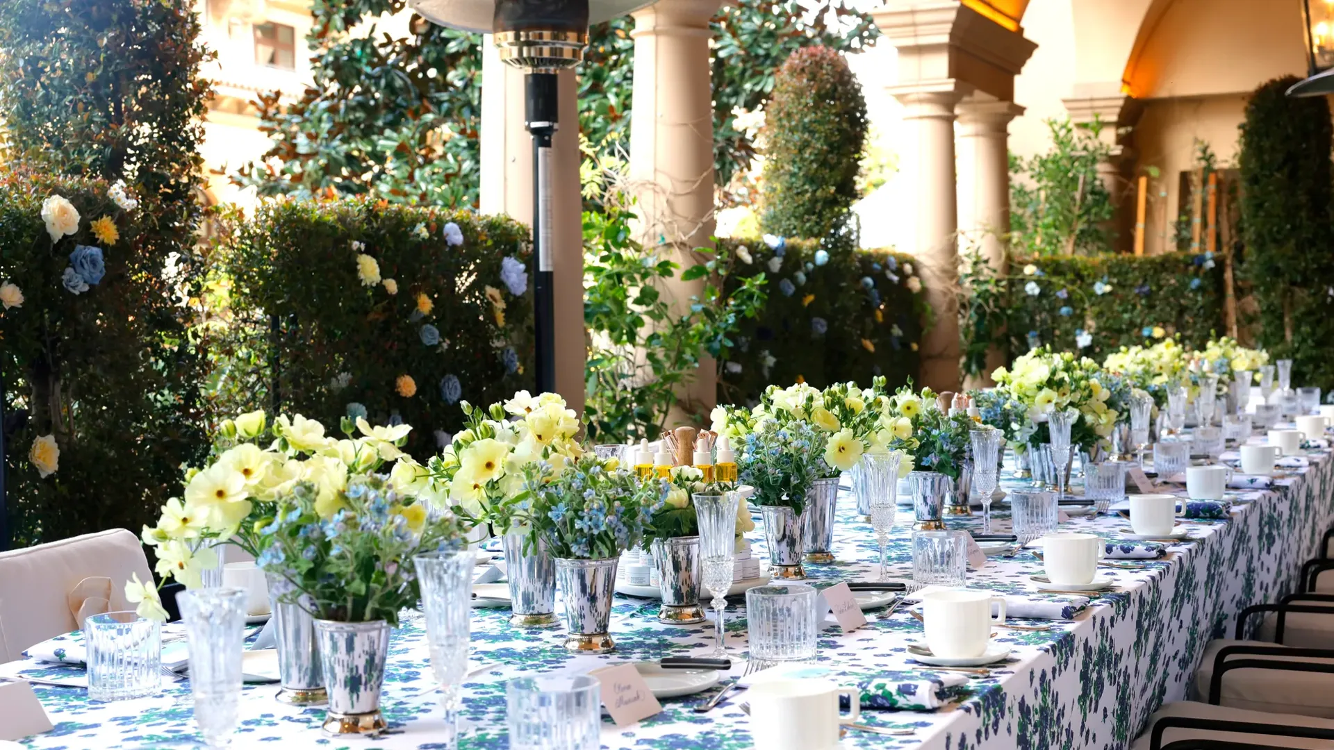 Kitchen Patio long table dressed in blue-floral linens with pastel centerpieces beneath an arched colonnade and garden hedges.