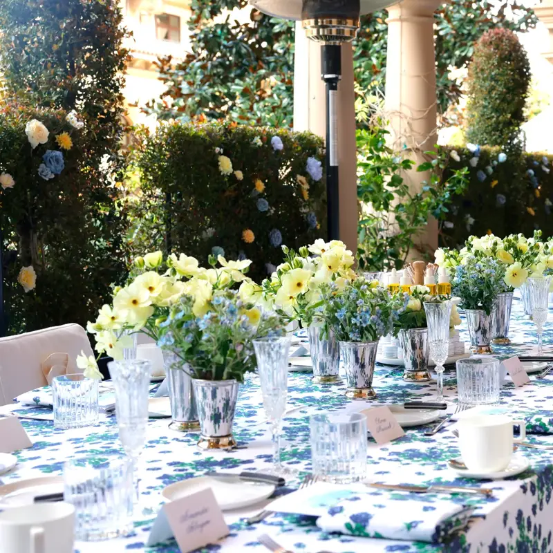 Kitchen Patio long table dressed in blue-floral linens with pastel centerpieces beneath an arched colonnade and garden hedges.
