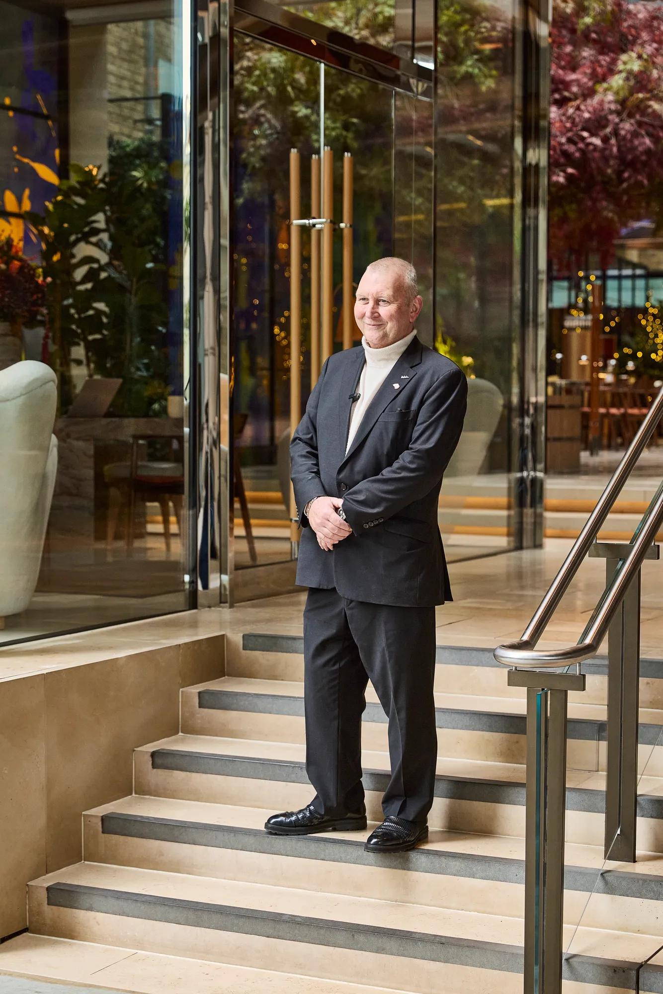 A man in a dark suit stands on steps outside a modern glass building.