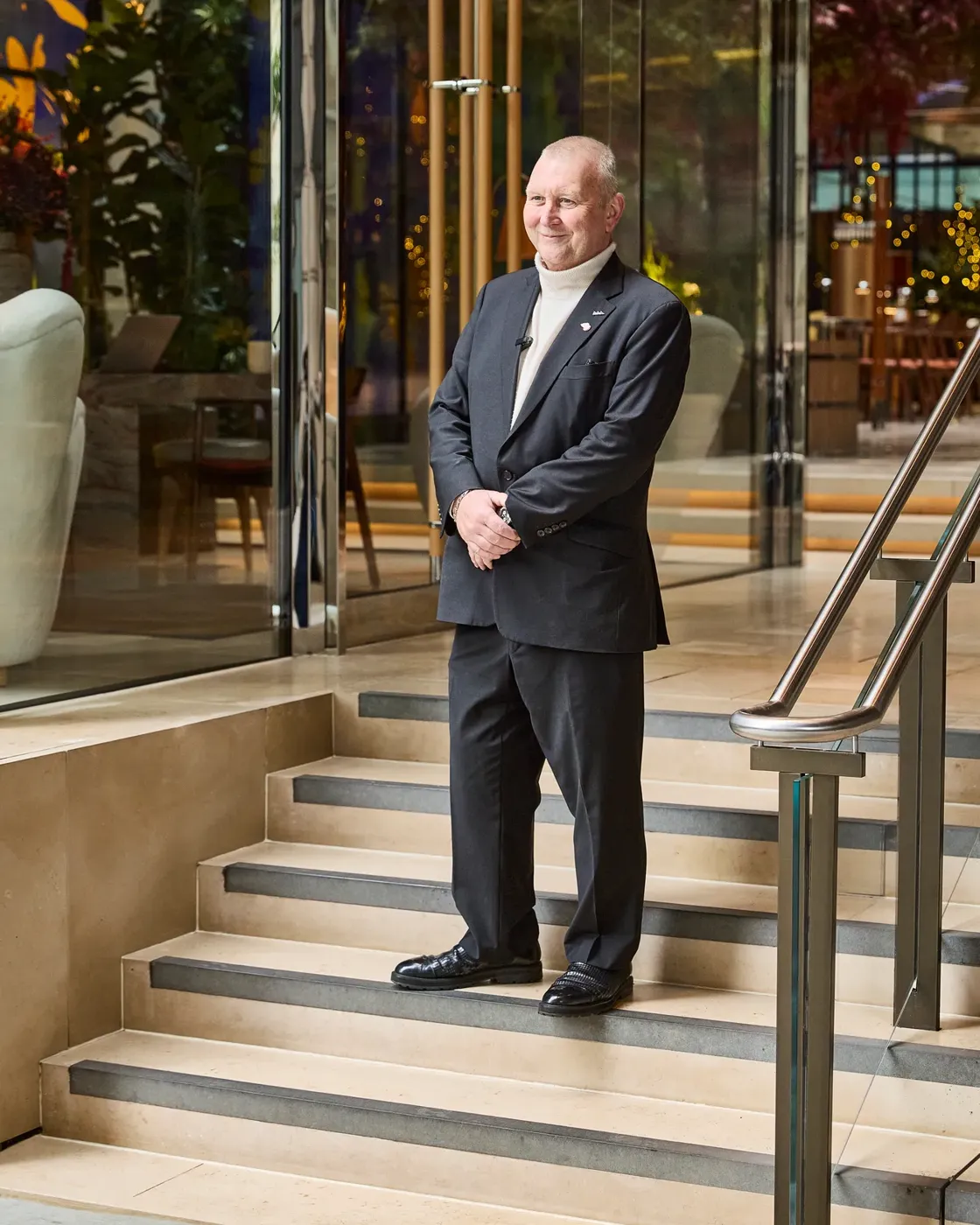 A man in a dark suit stands on steps outside a modern glass building.